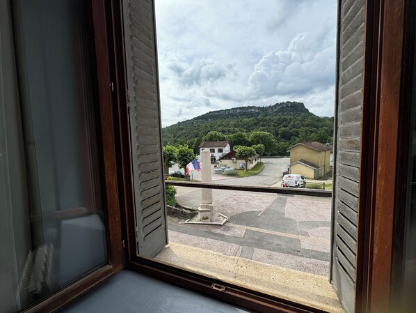 A view from a window with a wooden frame, overlooking a parking area, a small building with a flag, and a hill in the background.