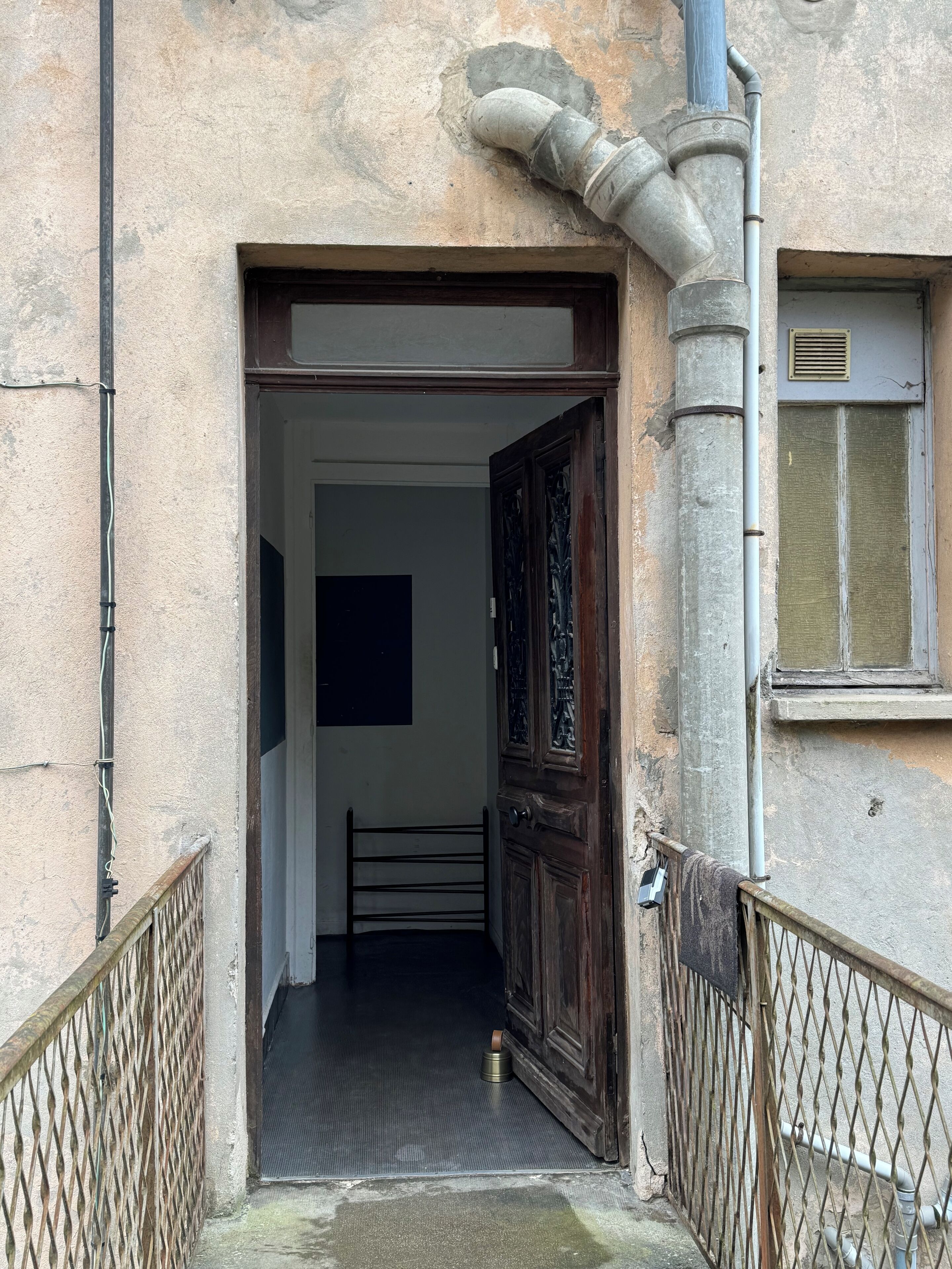 Un ingresso stretto con un cancello in metallo, una porta in legno finemente intagliata e un piccolo contenitore metallico a terra.