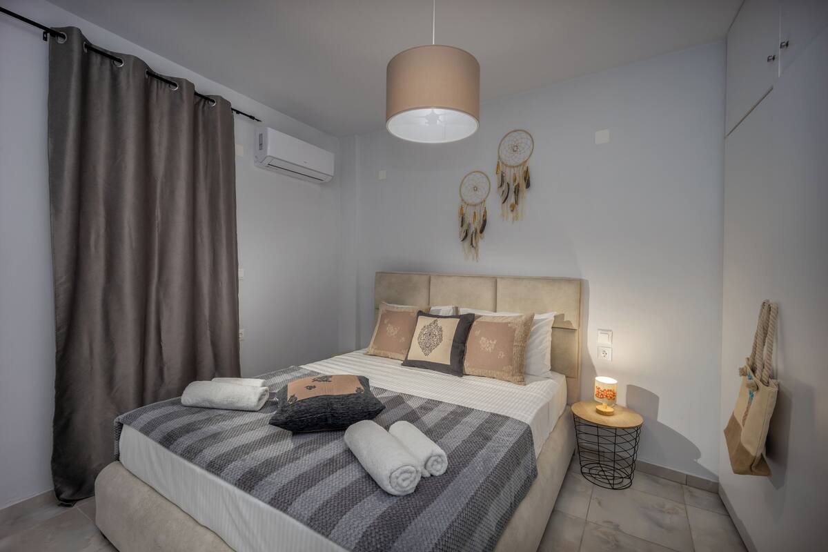 Una cama bien hecha con una manta a rayas, almohadas y toallas enrolladas. Hay una lámpara de noche, un perchero de pared y una decoración de atrapasueños.