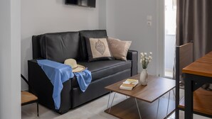 A modern living room with a black leather sofa, a wooden coffee table, a flat-screen TV mounted on the wall, and a view of the outdoors through a sliding door. - Sun City Apartments by Estia (Agios Nikolaos)