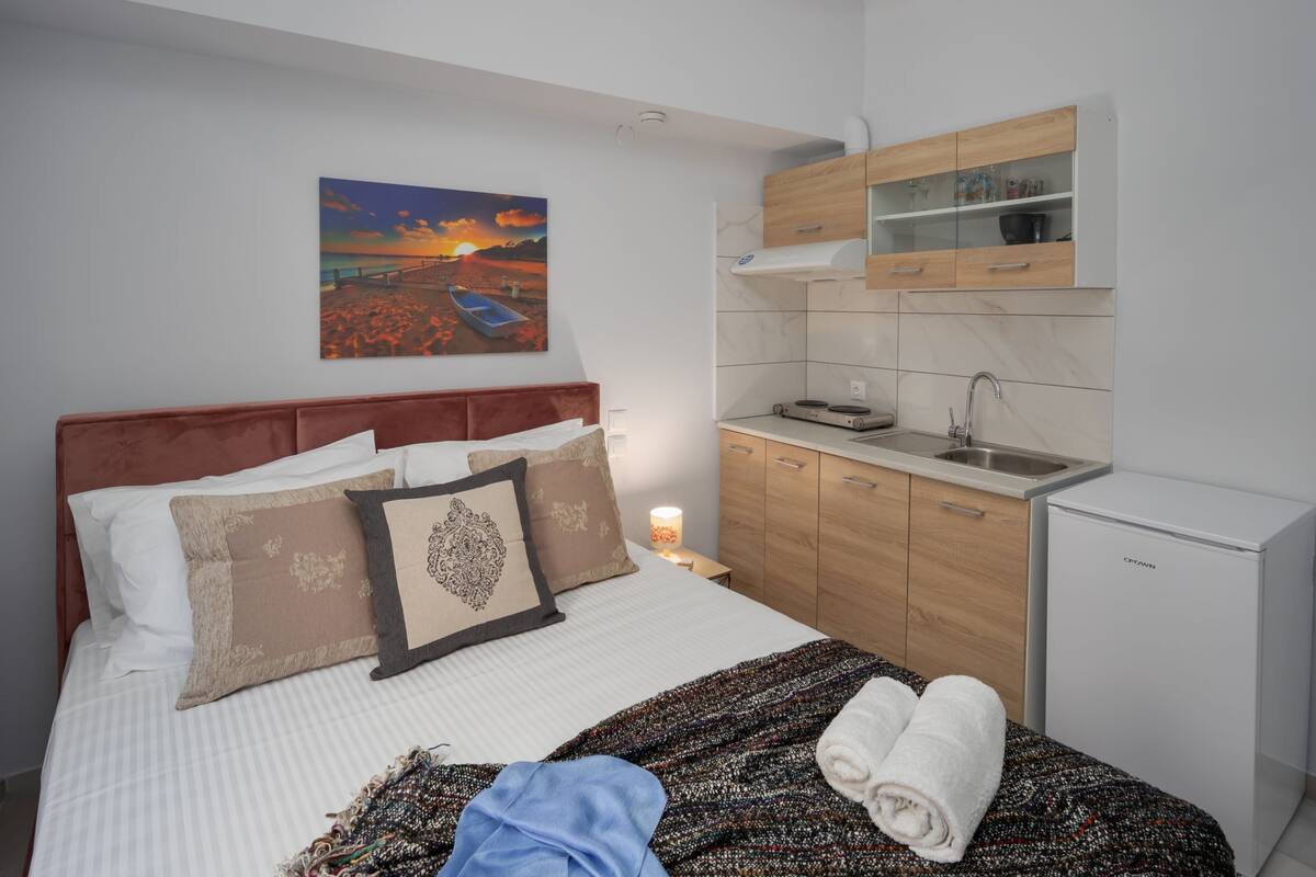 Un dormitorio con cama, almohadas, manta, un cuadro, una mesita de noche y una pequeña cocina americana con refrigerador.