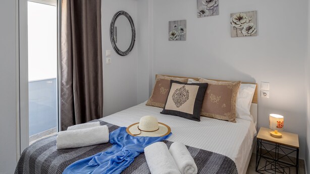 A neatly made bed with a blue towel, a straw hat, and rolled towels. There are floral wall art pieces and a small table with a lamp beside the bed. - Sun City Apartments by Estia (Agios Nikolaos)