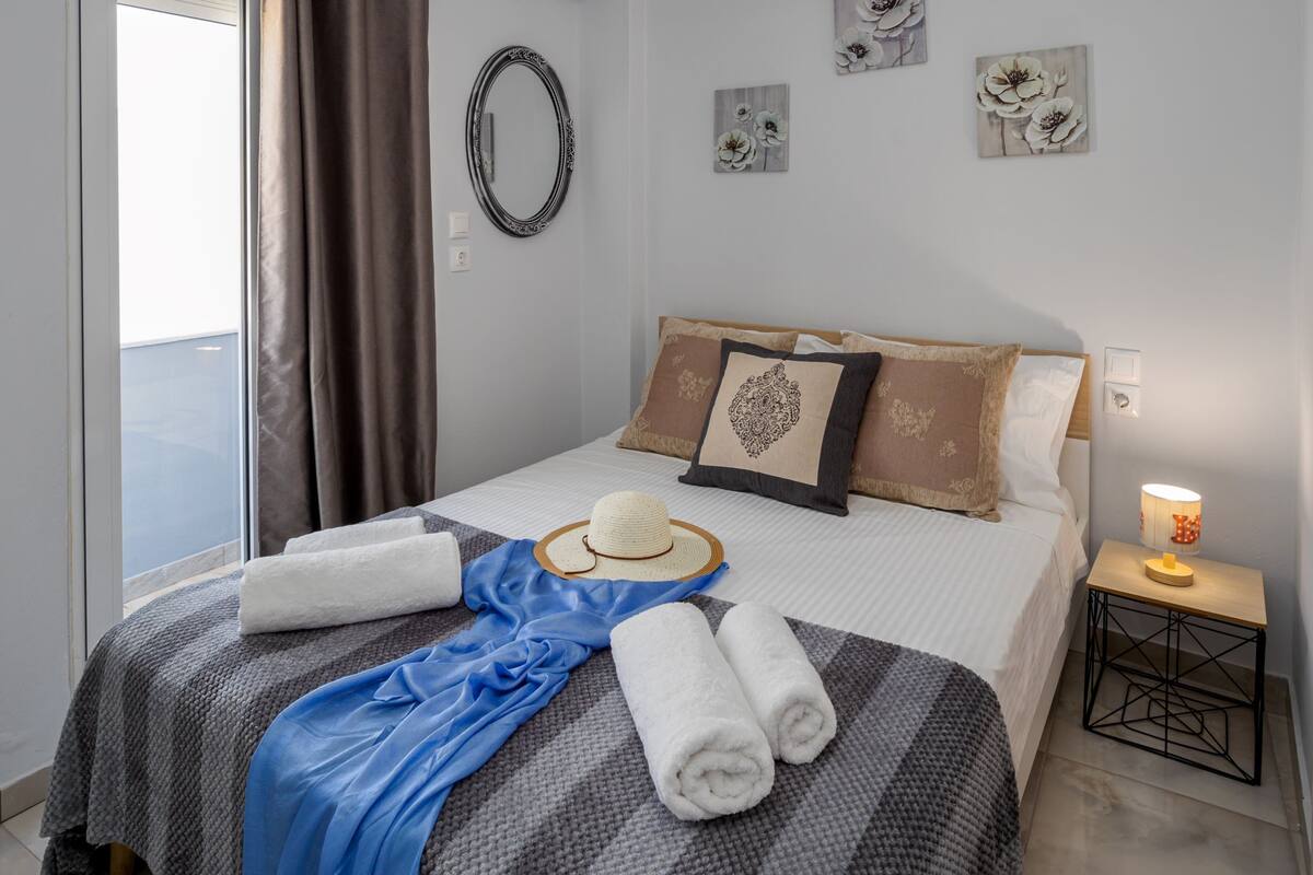 Una cama hecha con cuidado, una toalla azul, un sombrero de paja y toallas enrolladas. Se observan detalles decorativos de flores en la pared y una mesita de noche con una lámpara junto a la cama.
