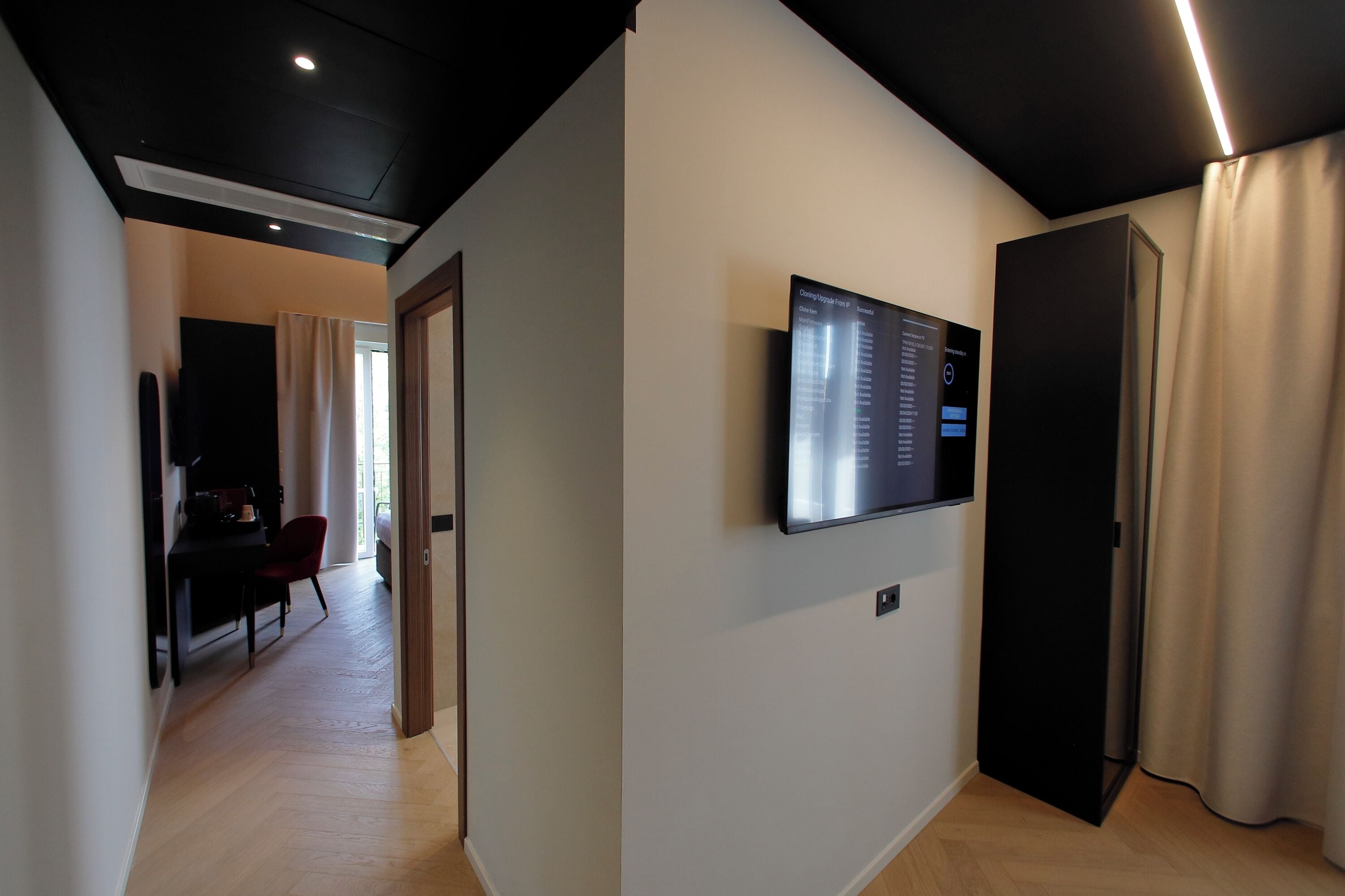 A modern hotel room with a flat-screen TV mounted on the wall, a wooden floor, and a door with a curtain.