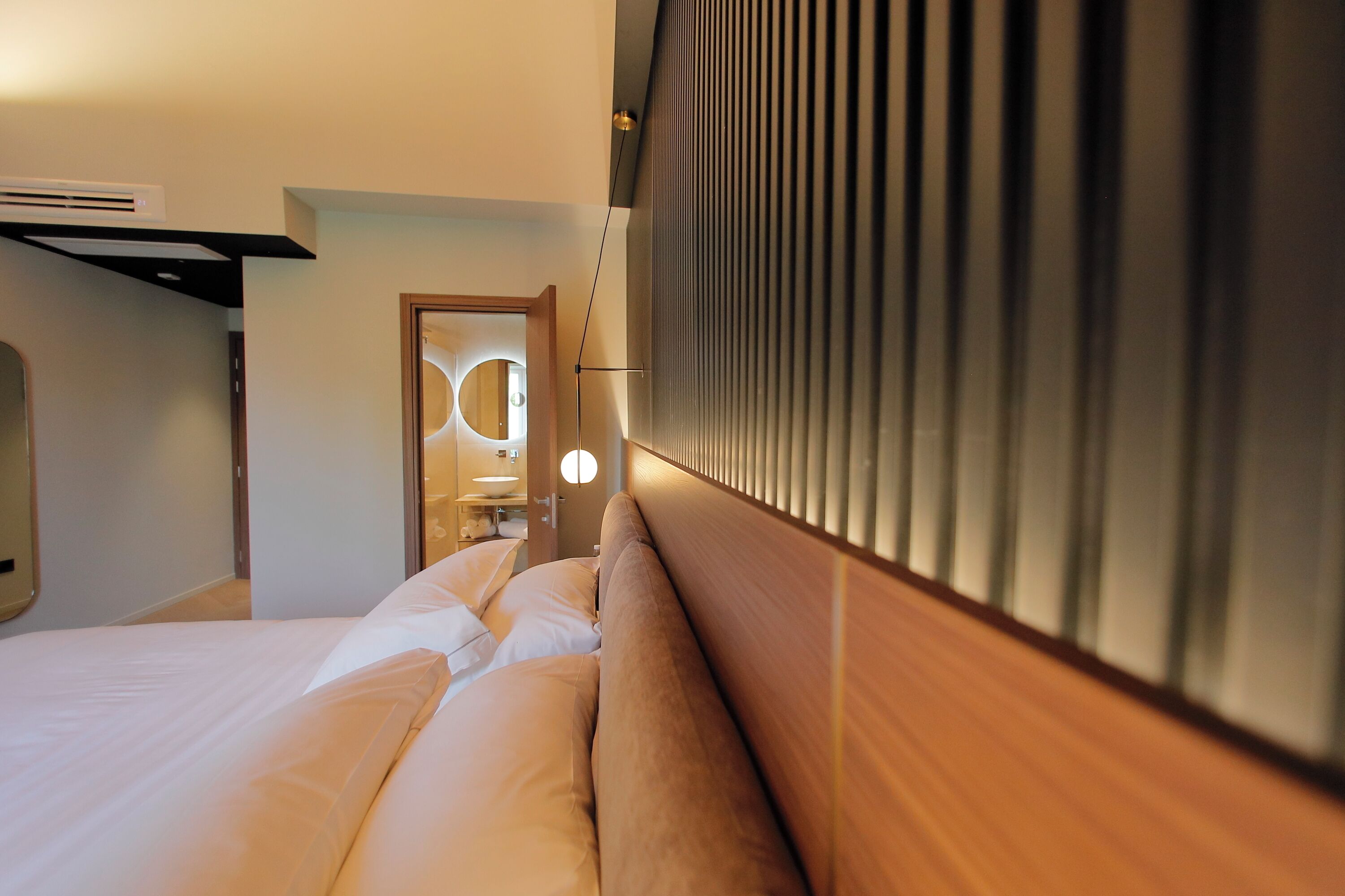 A hotel room with a bed, a headboard with vertical slats, a wall-mounted air conditioning unit, and a door leading to another room.