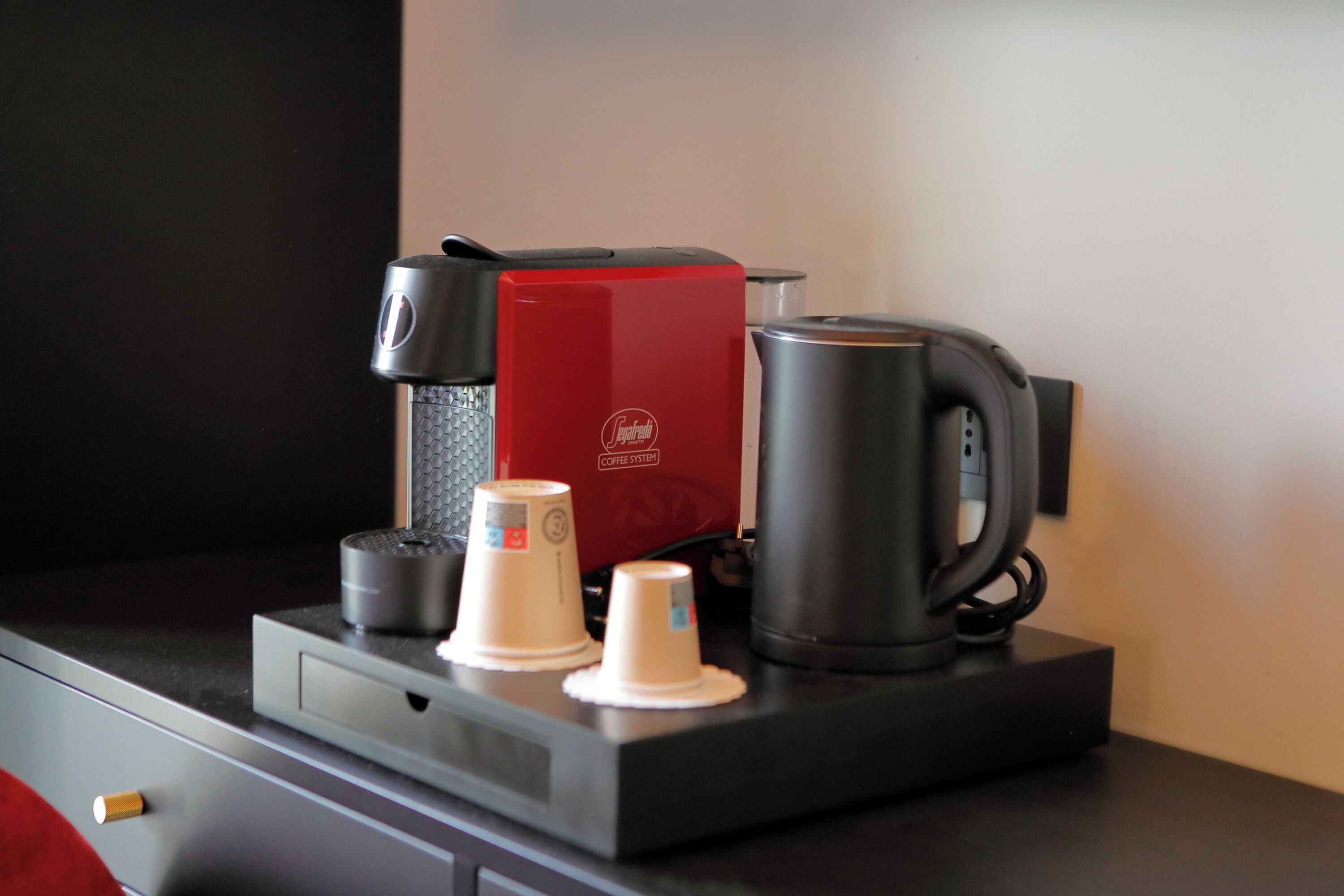Una estación de café con una cafetera roja, dos vasos de papel y una taza negra sobre una encimera.