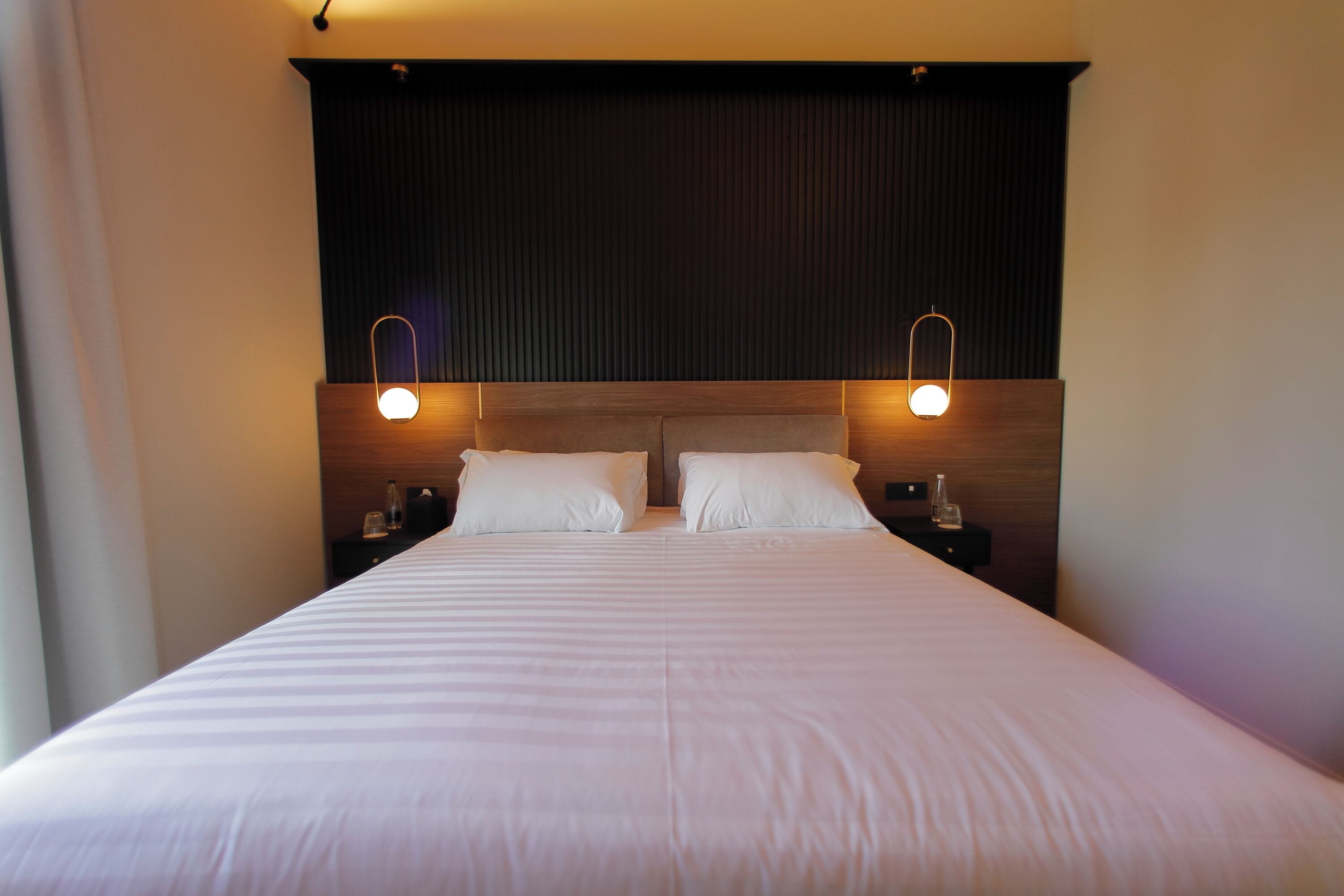 Una camera d'albergo con un letto grande, due comodini e una testiera scura.