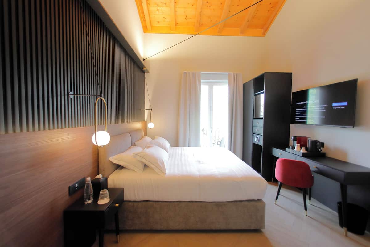 A modern hotel room with a large bed, a desk with a chair, a TV mounted on the wall, and a window with curtains.