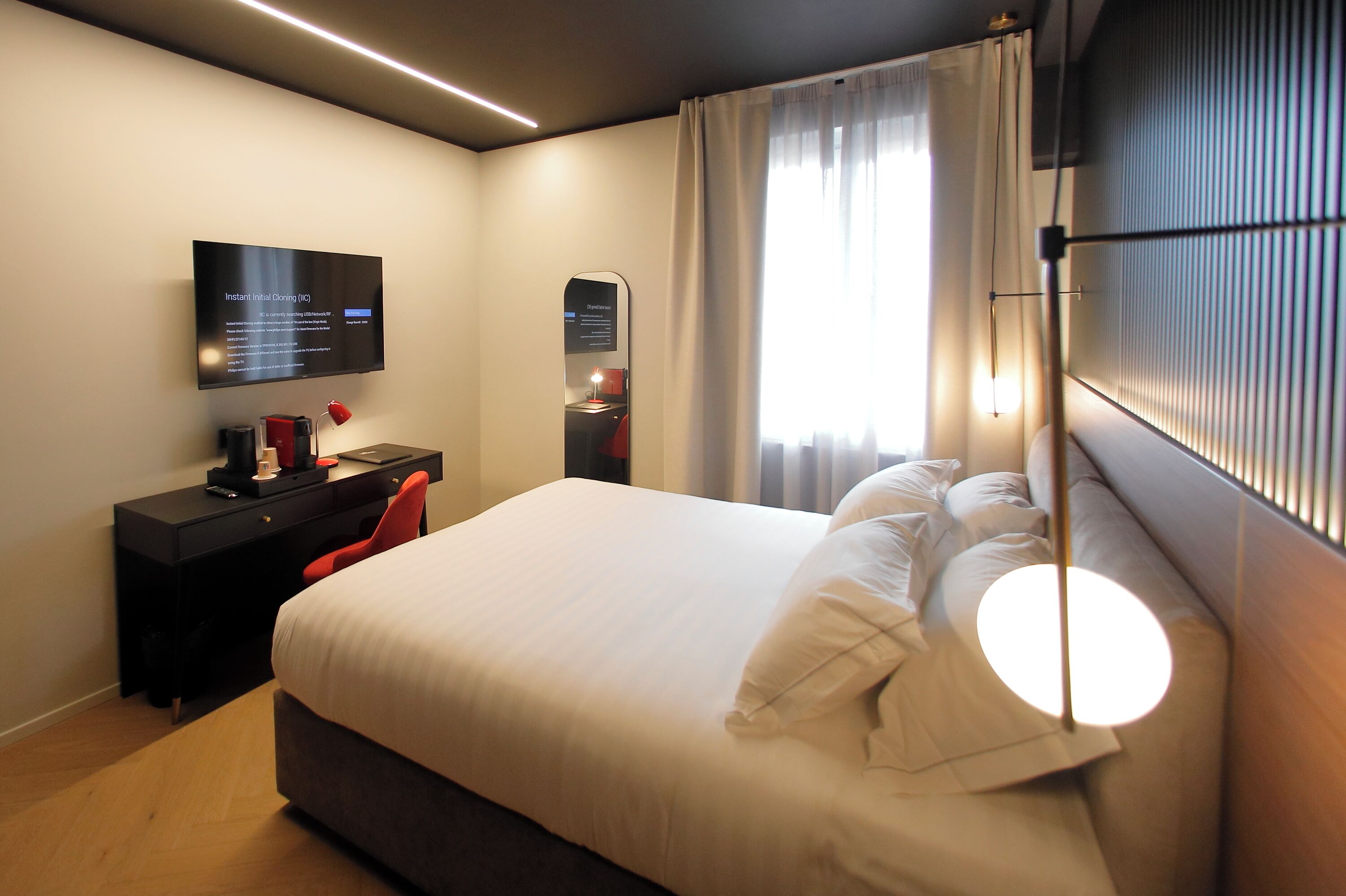 A modern hotel room with a large bed, a desk with a computer, a flat-screen TV, and a large window with curtains.