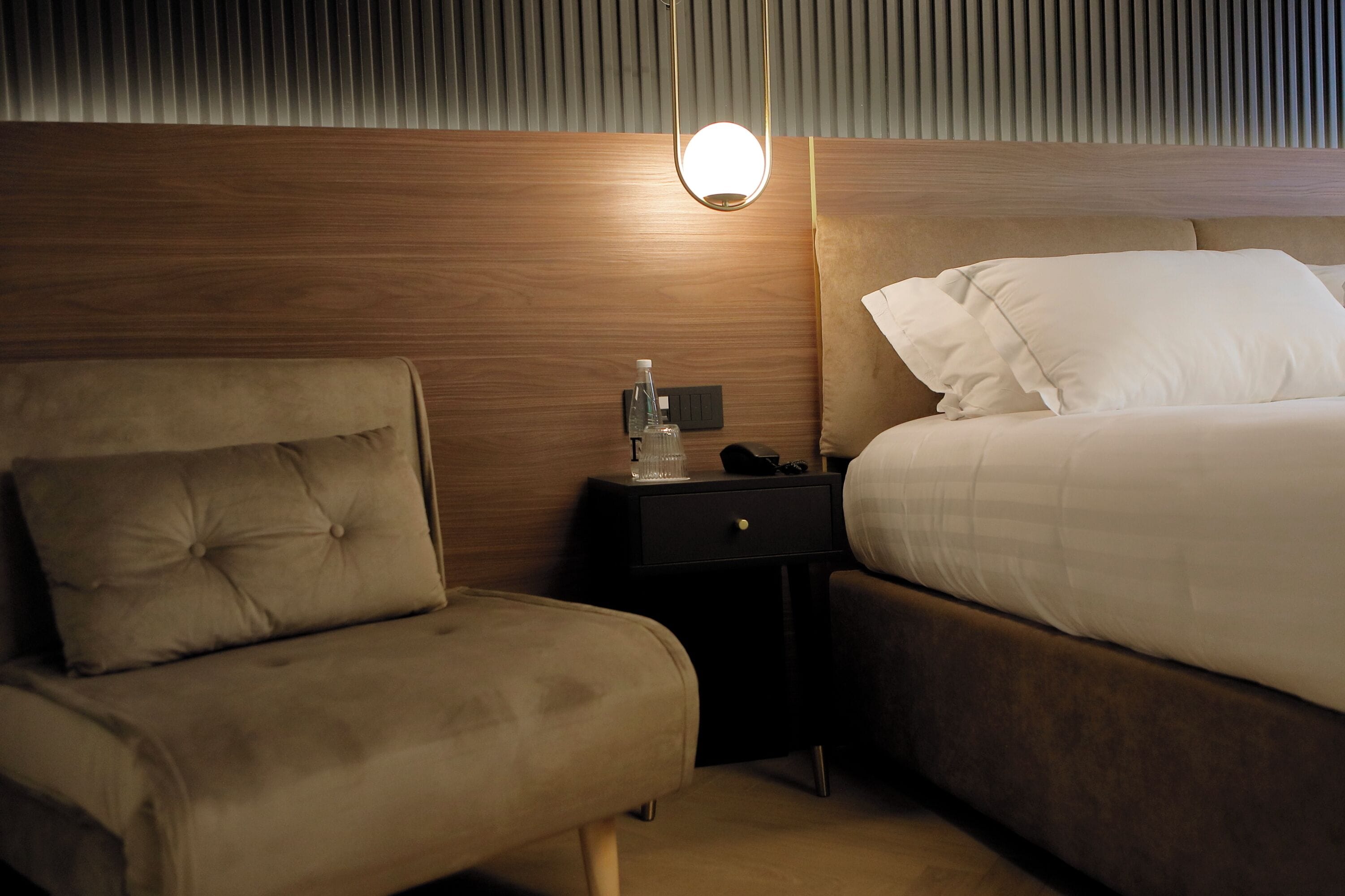 A hotel room with a wooden headboard, a bedside table with a lamp, a chair, and a bed with white bedding.