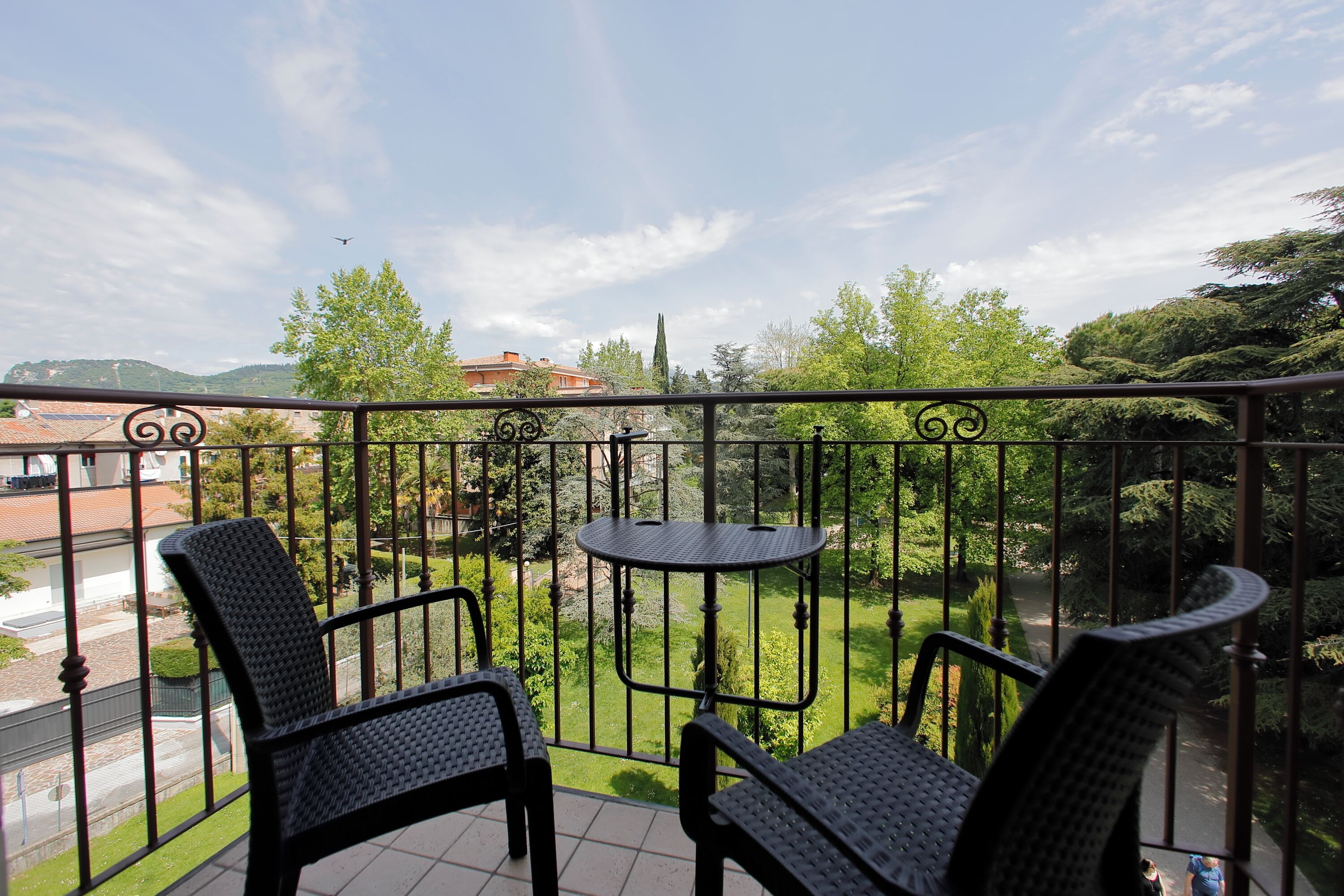 Un balcon avec deux fauteuils en osier et une petite table ronde, donnant sur un jardin avec des arbres et un bâtiment situé au loin.