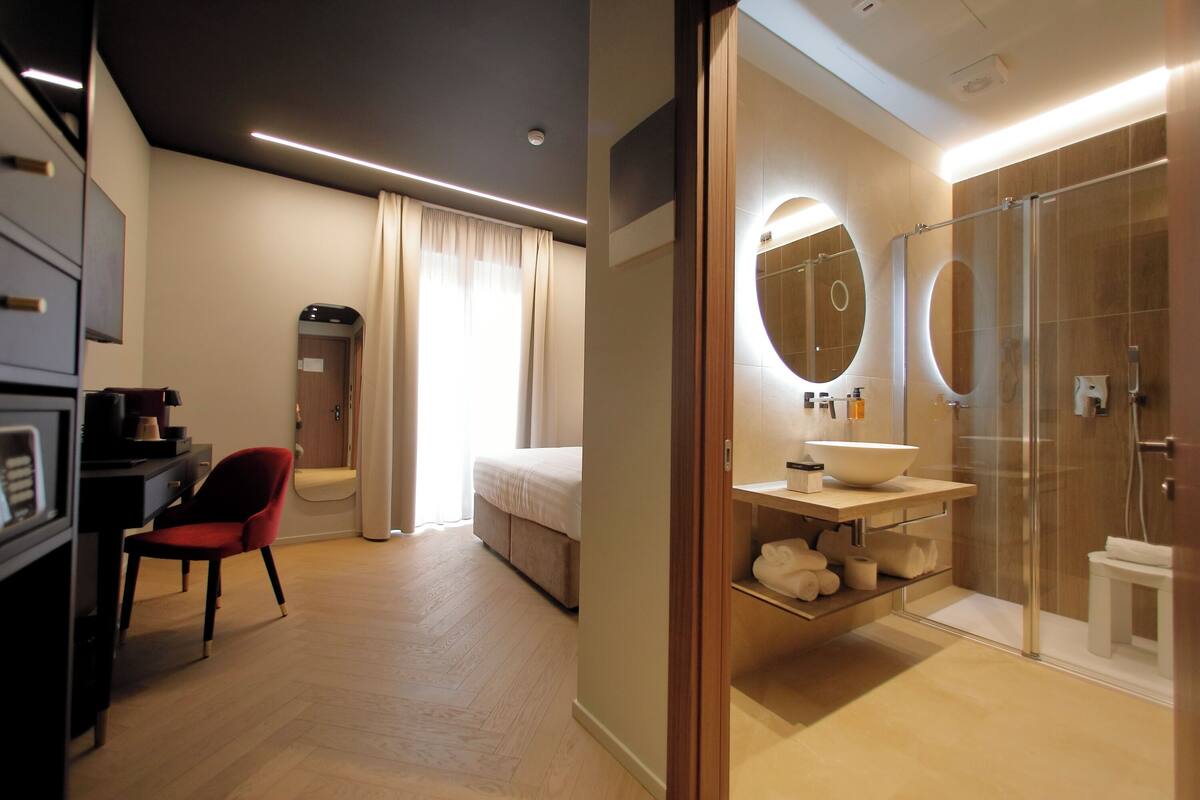 Une chambre d’hôtel moderne équipée d’un lit, d’un bureau, d’une chaise rouge, d’une armoire et d’une salle de bain avec une douche en verre et un grand miroir.