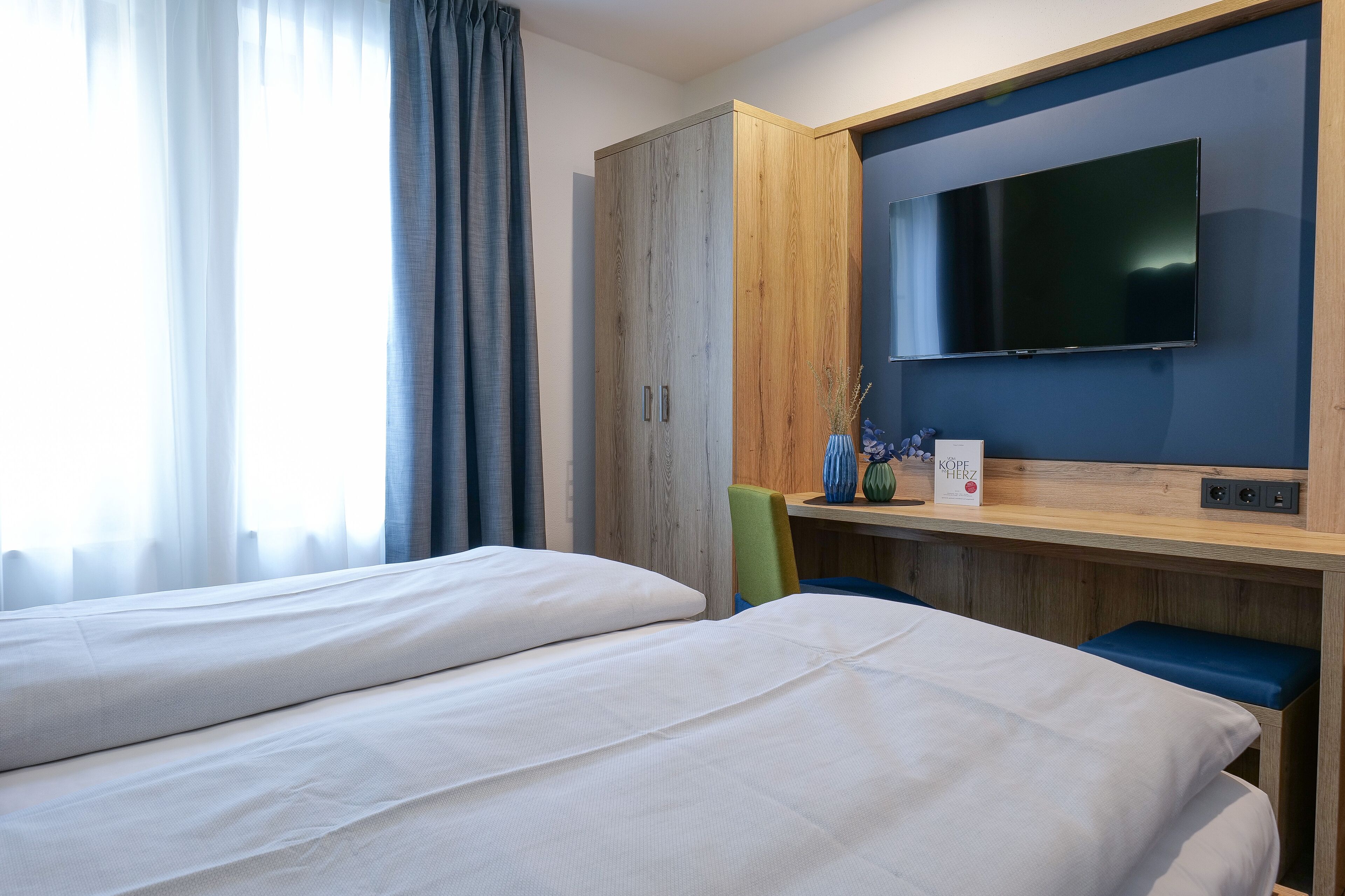 A hotel room with two beds, a wooden wardrobe, a flat-screen TV, and a small table with a vase.