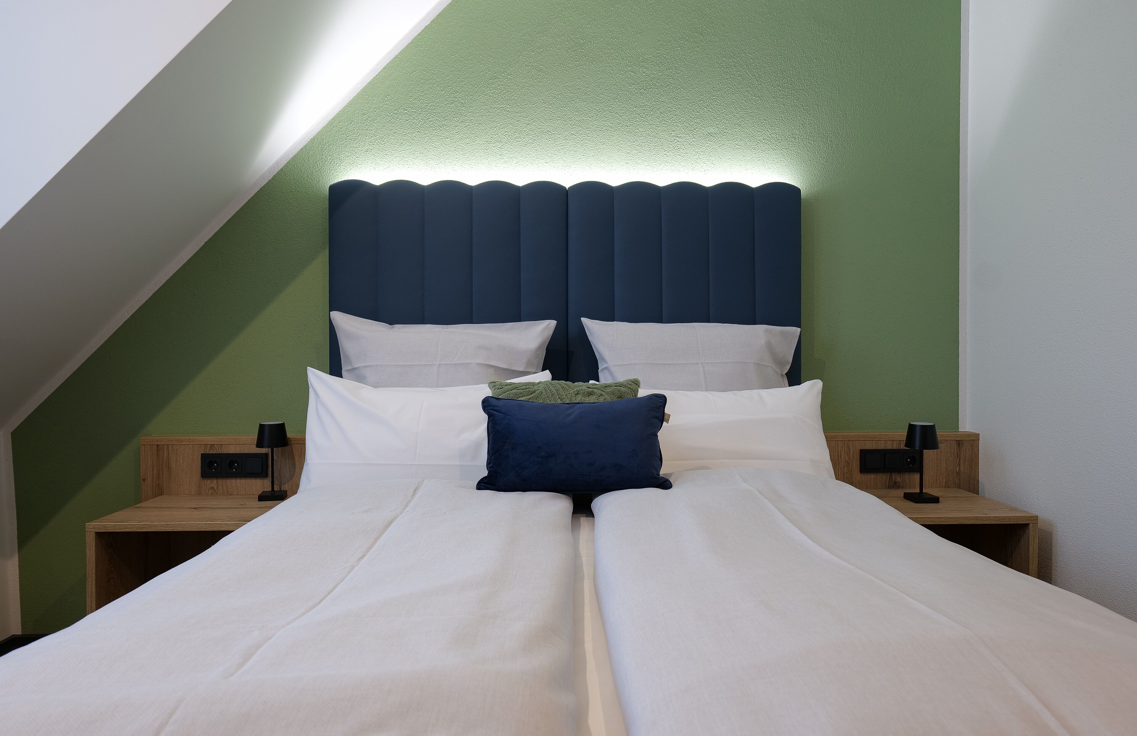 A double bed with white and blue bedding, two bedside tables with lamps, and a green accent wall.