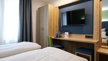 Habitación de hotel con dos camas, un armario de madera, un televisor montado en la pared y un escritorio pequeño con un jarrón y una caja.