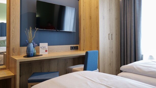 A hotel room with a bed, a desk with a flat-screen TV, a wooden wardrobe, and a blue chair. - Reos Hotel Isny (Isny im Allgaeu)