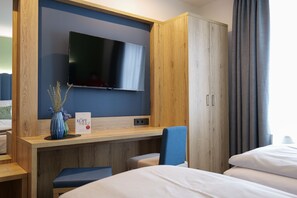 Quarto de hotel com cama, escrivaninha com televisão de tela plana, guarda-roupa de madeira e uma cadeira azul.