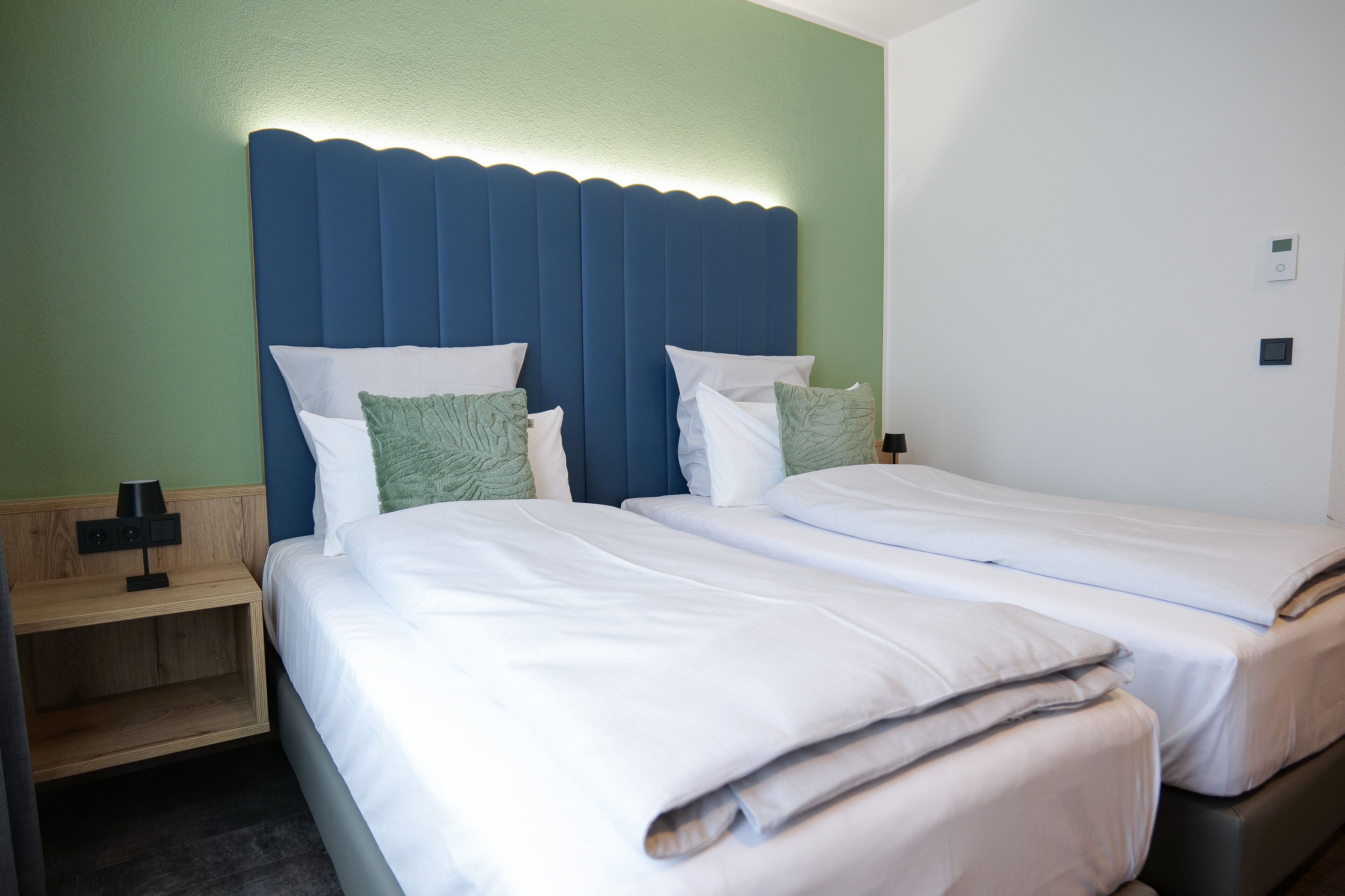 Habitación de hotel con dos camas individuales, una mesita de noche de madera con lámpara y una pared decorativa verde.