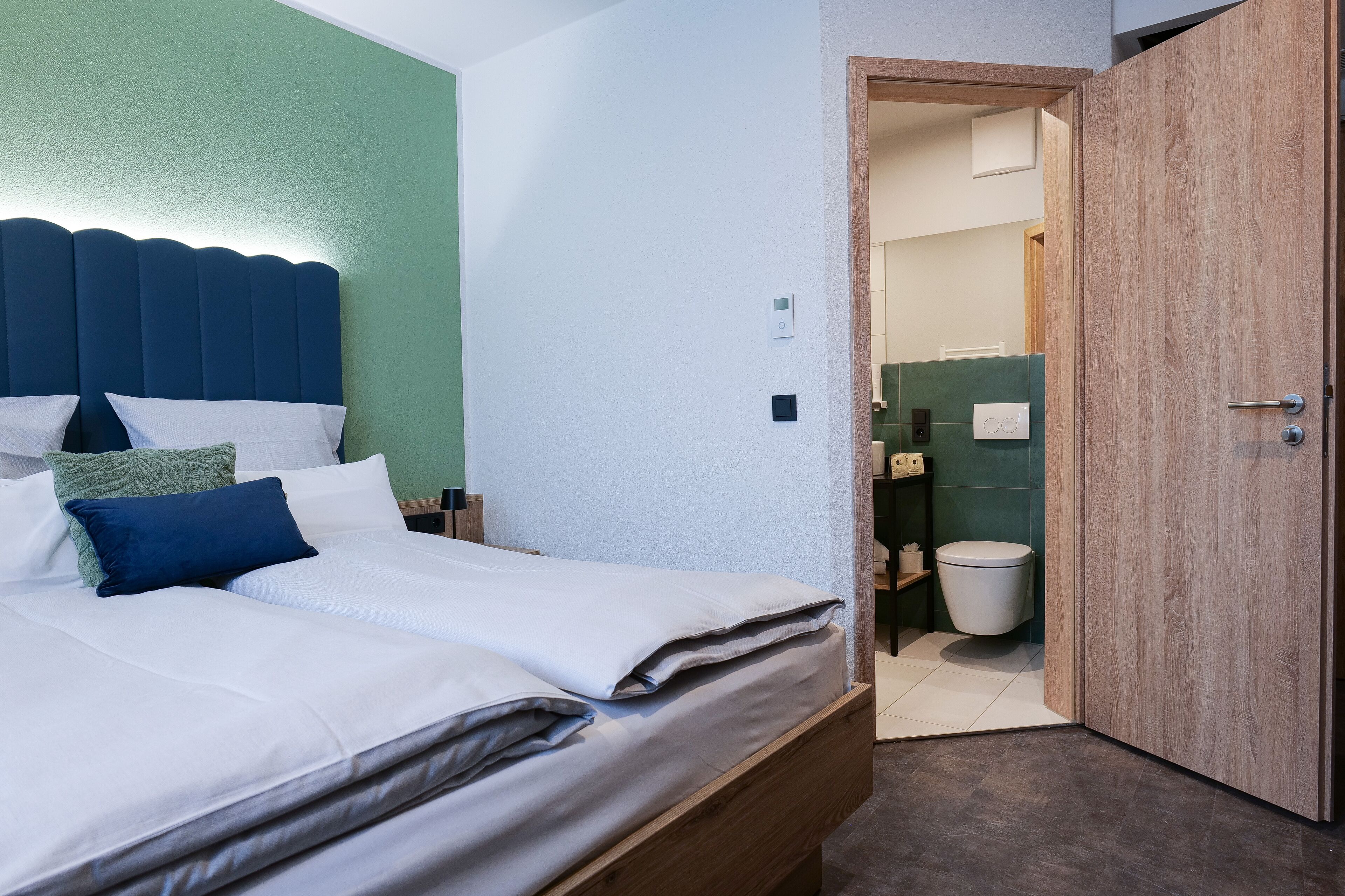Een net opgemaakt bed met wit- en blauwe beddengoed, een houten hoofdbord en een groene accentmuur.