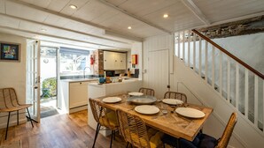 A dining area with a wooden table set for four, surrounded by wicker chairs, a kitchen in the background, and a staircase with white railings. - Sea Horses (Port Isaac)