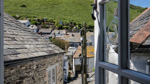 A narrow street with stone buildings, a hill in the background, and a view through a window with a metal frame. - Sea Horses (Port Isaac)