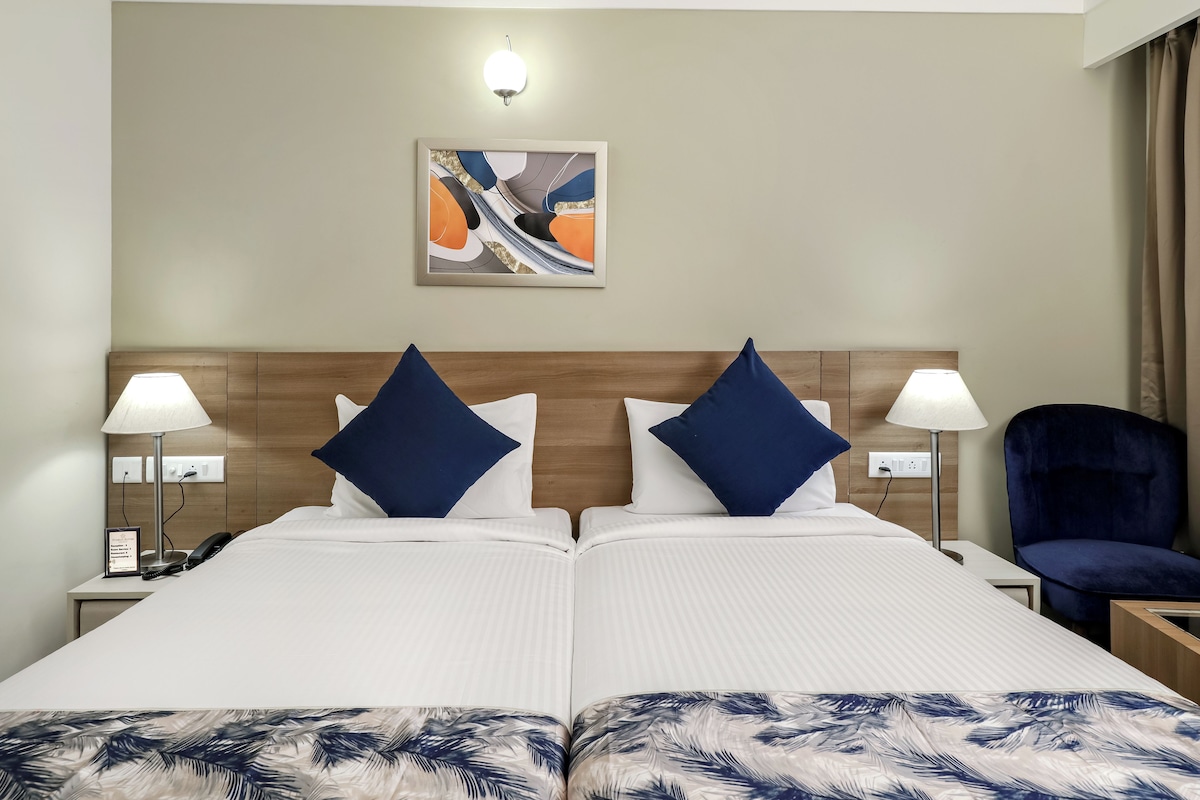 A hotel room with a double bed, two bedside lamps, a nightstand, a chair, a painting on the wall, and a power outlet.