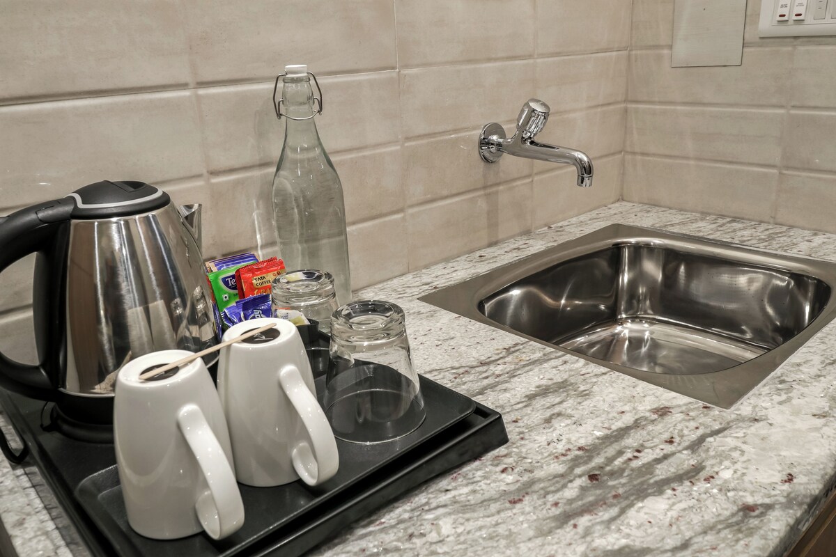 Uma cozinha com uma chaleira de aço inoxidável, duas canecas brancas, uma garrafa de vidro e uma caixa de saquinhos de chá sobre uma bancada de mármore.