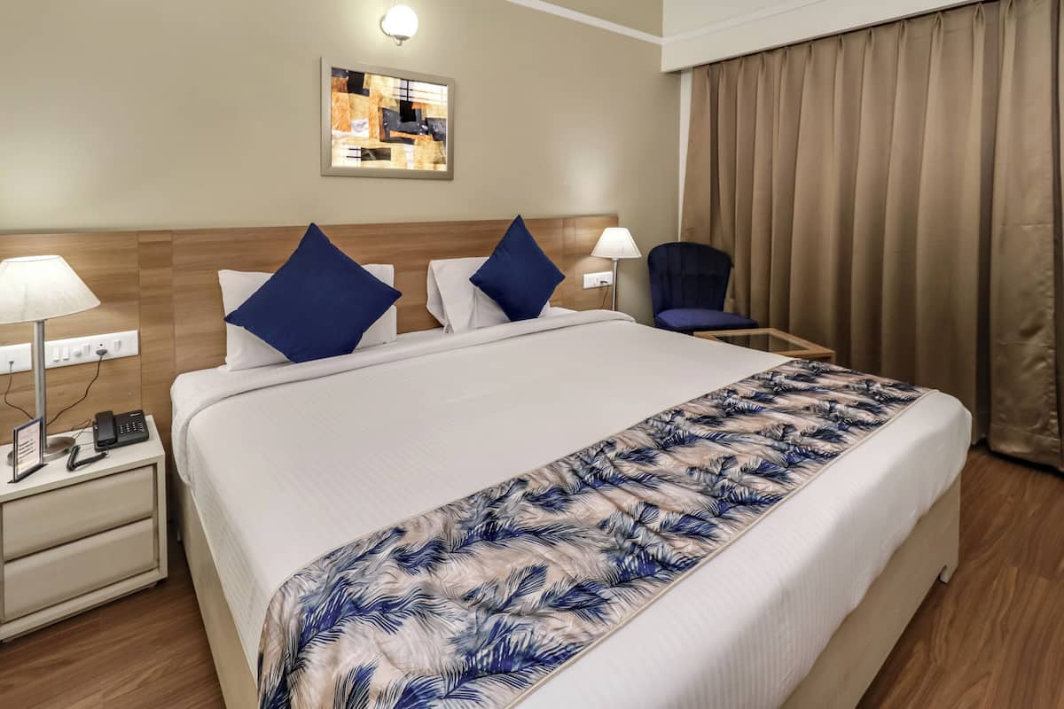 Ett hotellrum med en stor säng, tv å sänglampor, en stol, ett skrivbord med en telefon och en lampa samt en gardin.