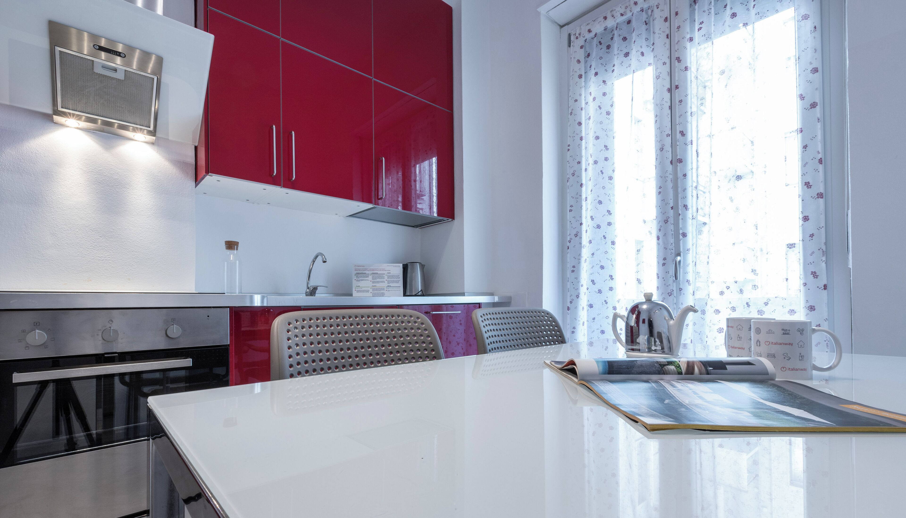 A modern kitchen with red cabinets, a white countertop, and a window with floral curtains.