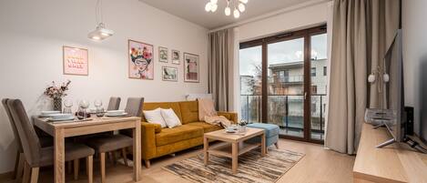 A modern living room with a dining area, a sofa, a coffee table, and a TV. There are curtains, a window, and artwork on the walls.