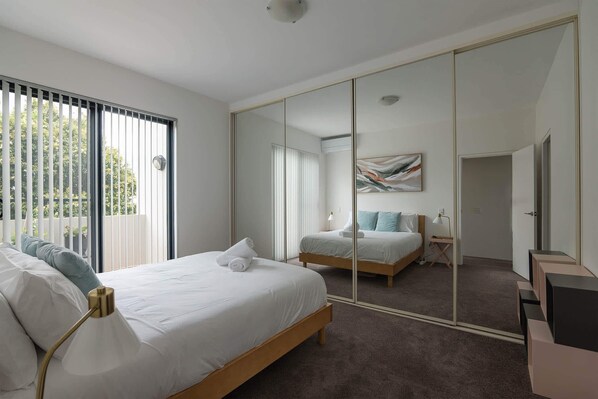 A modern bedroom with a large mirror, a bed with white linens, a bedside table with a lamp, and a view through the window. - Treetops Camperdown, Inner City Bliss w Parking (Camperdown)
