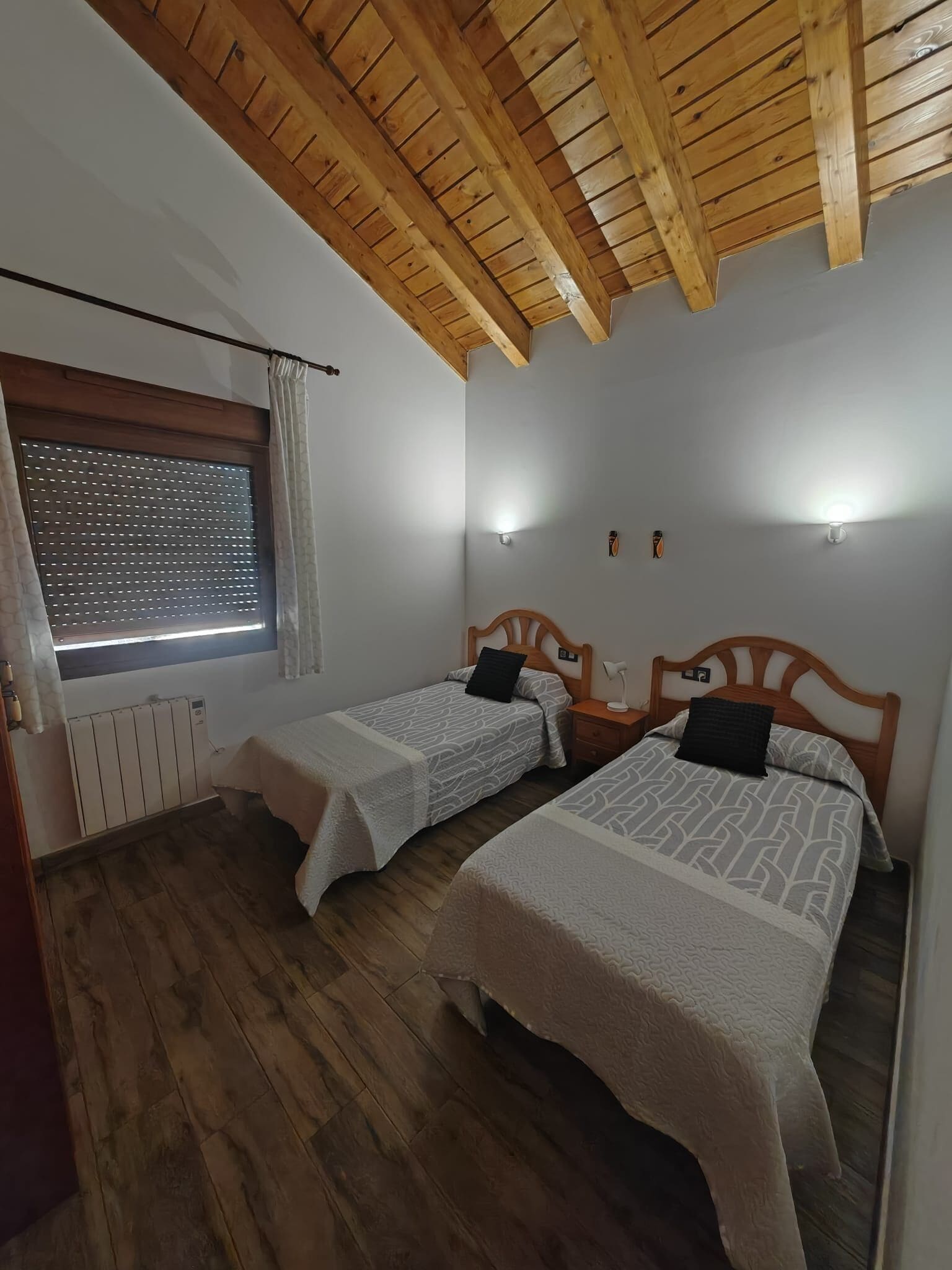 A bedroom with two beds, wooden ceiling beams, a window with blinds, and a radiator.