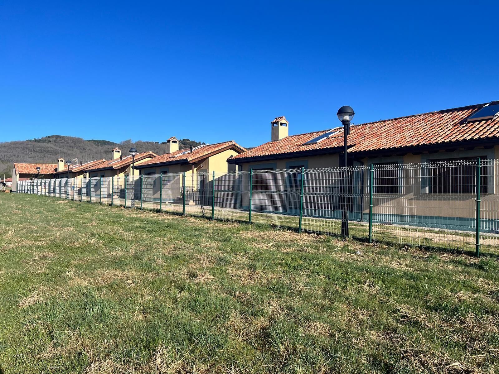 A row of houses with a metal fence in front, a clear blue sky, and a grassy field.
