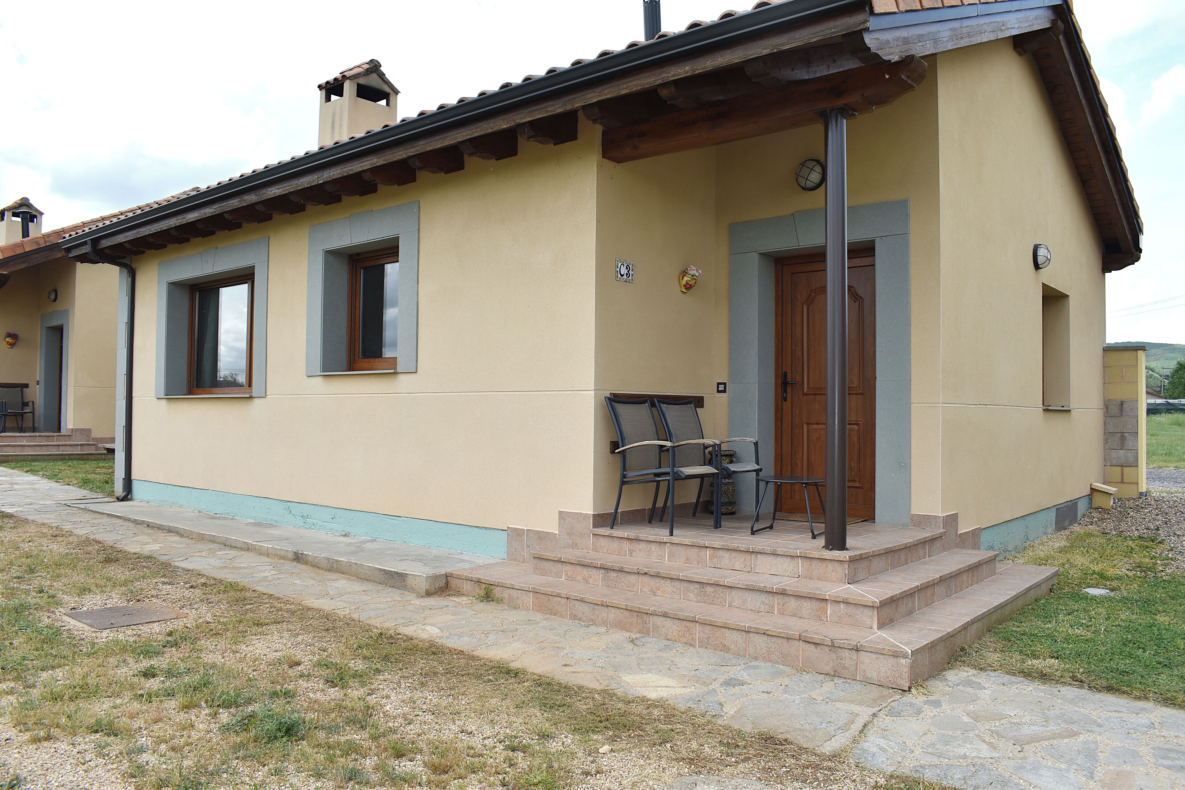 A two-story building with a beige facade, a tiled roof, and a small porch with a table and chairs.