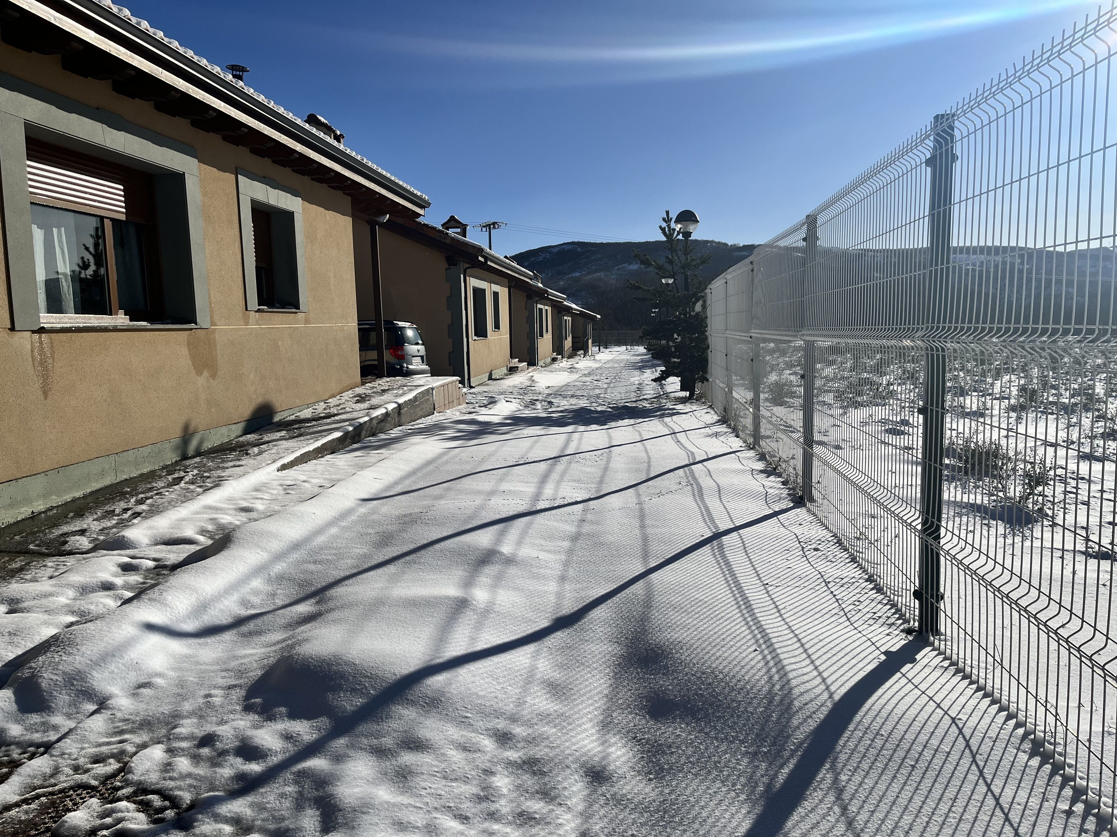 A snow-covered street with buildings on one side and a fenced area on the other.
