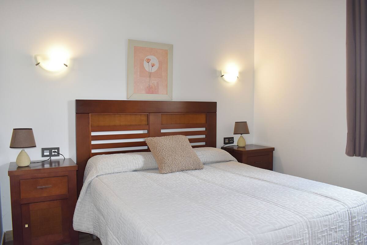 Un lit bien fait, avec une tête de lit en bois, deux tables de chevet avec des lampes et un luminaire fixé au mur.