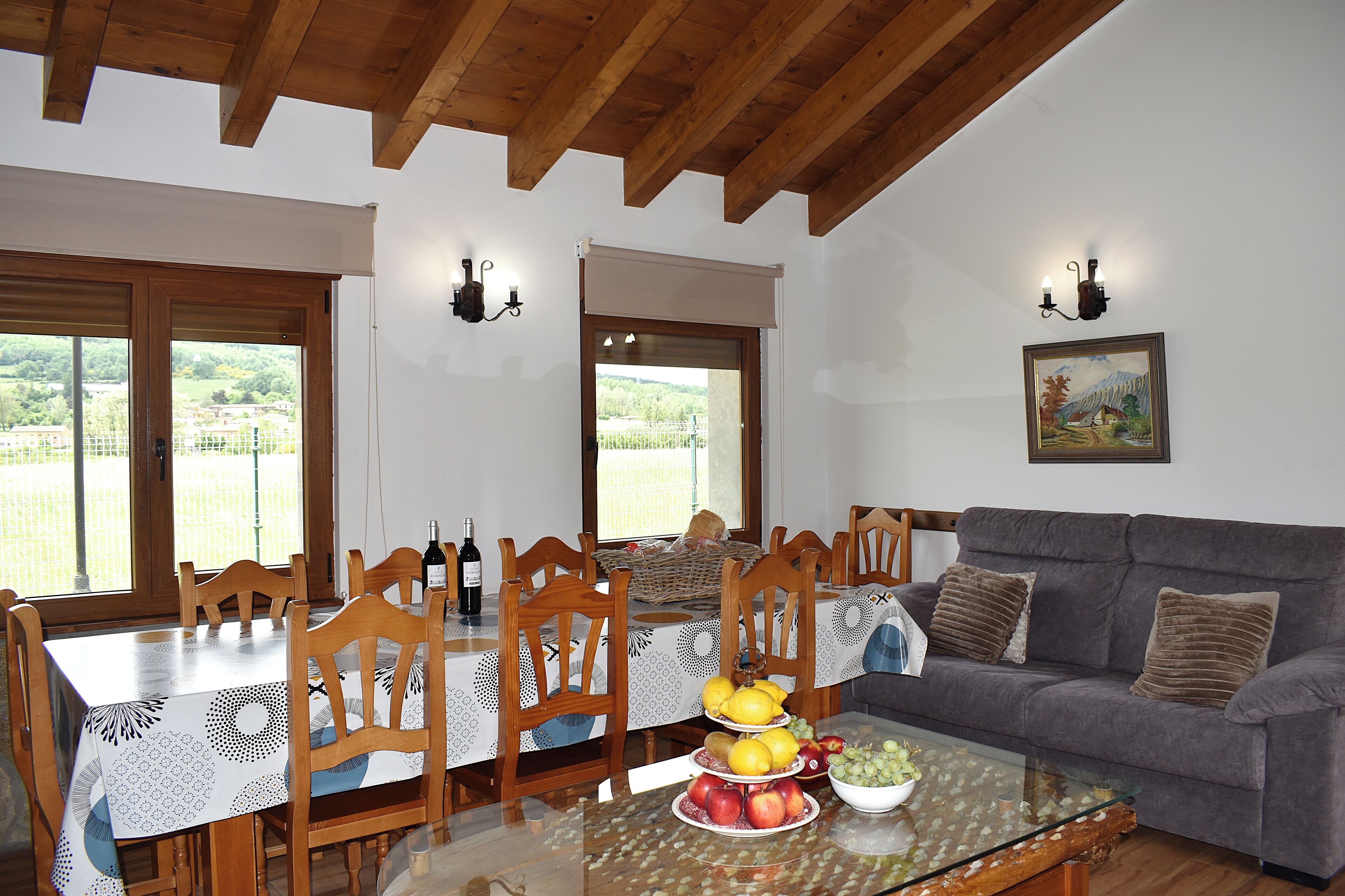 Un comedor con una mesa dispuesta para seis personas, rodeada de sillas de madera, una mesa de centro de vidrio con fruta, y un sofá gris.