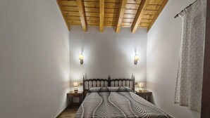 Une chambre avec un plafond en bois, deux lampes de chevet, un lit recouvert d’une couvre-lit à motifs et une fenêtre avec un rideau.