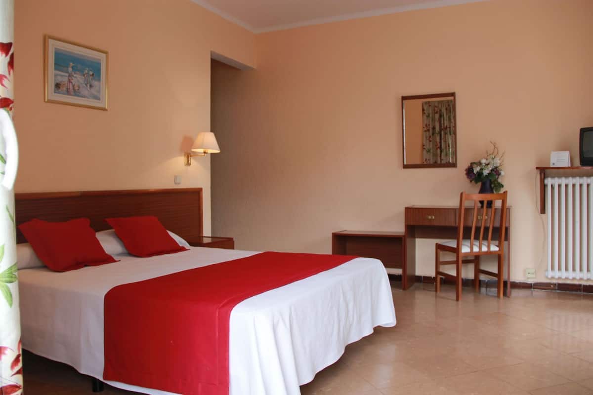 A hotel room with a bed, a desk, a chair, a lamp, and a radiator.