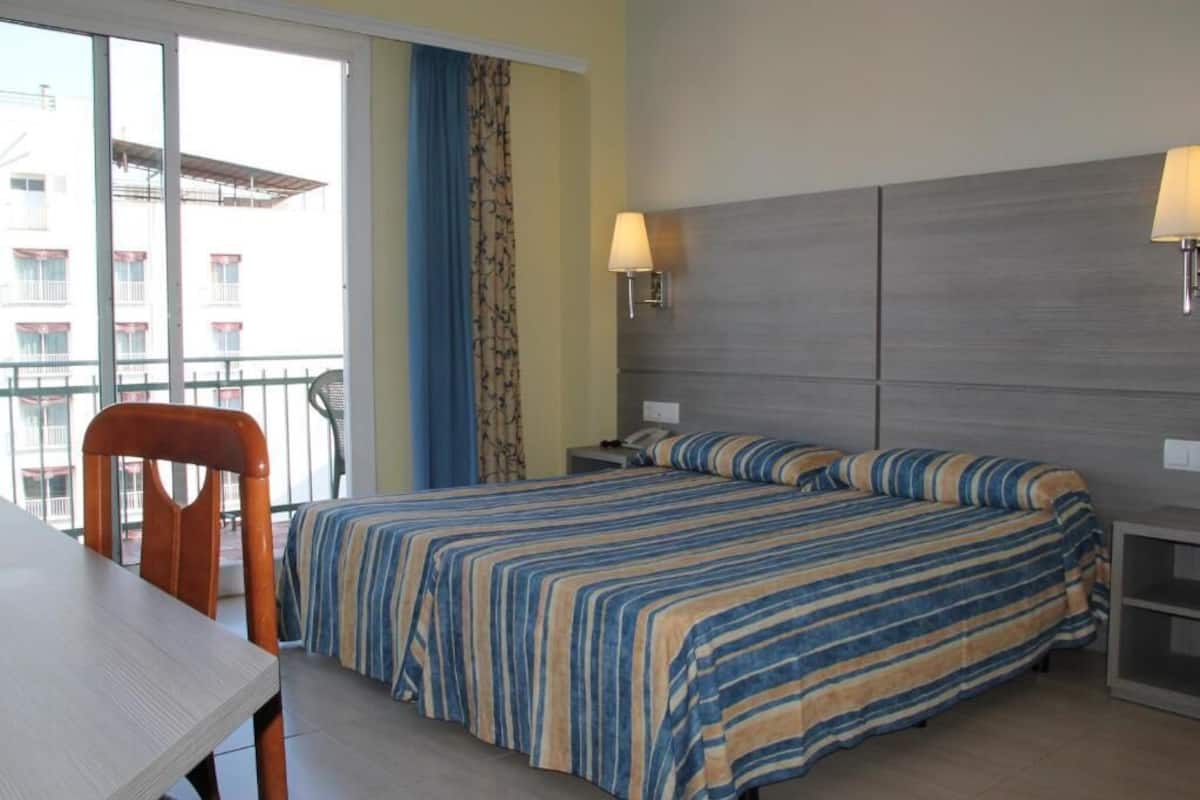 A hotel room with a bed, a desk, and a chair. There is a window with curtains, a lamp on the wall, and a view of buildings outside.