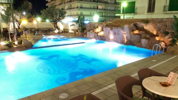 A swimming pool with a rock wall and a small waterfall feature. There are chairs and a table with a menu on the poolside. - Terramar (Calella)