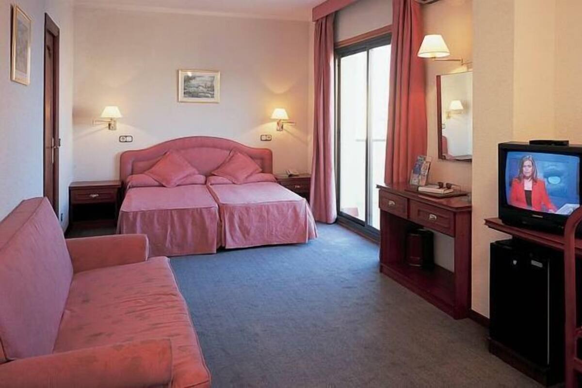 A hotel room with a double bed, a single bed, a desk with a television, a chair, and a sofa.