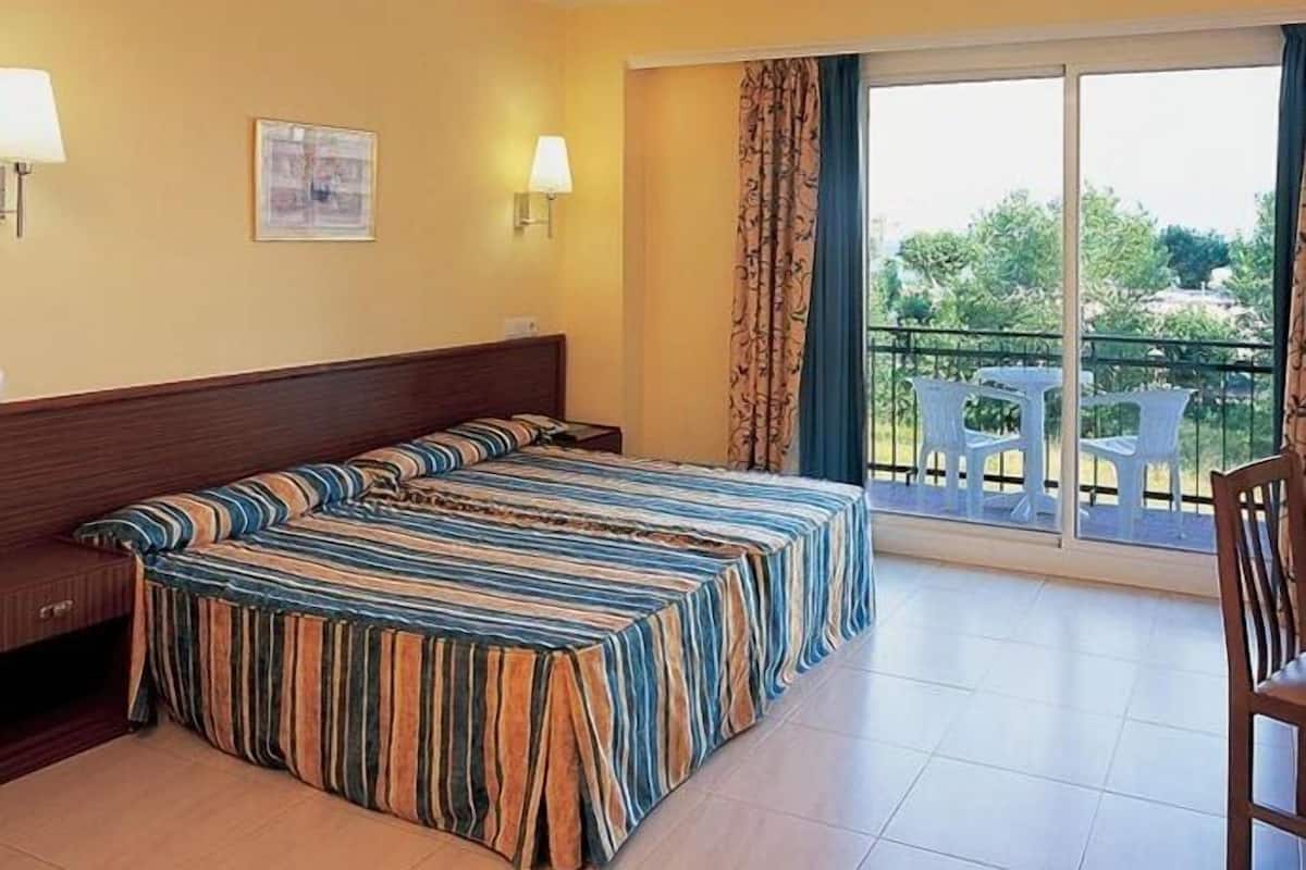 A hotel room with a bed, two bedside tables, a chair, a balcony with a view, and a window with curtains.