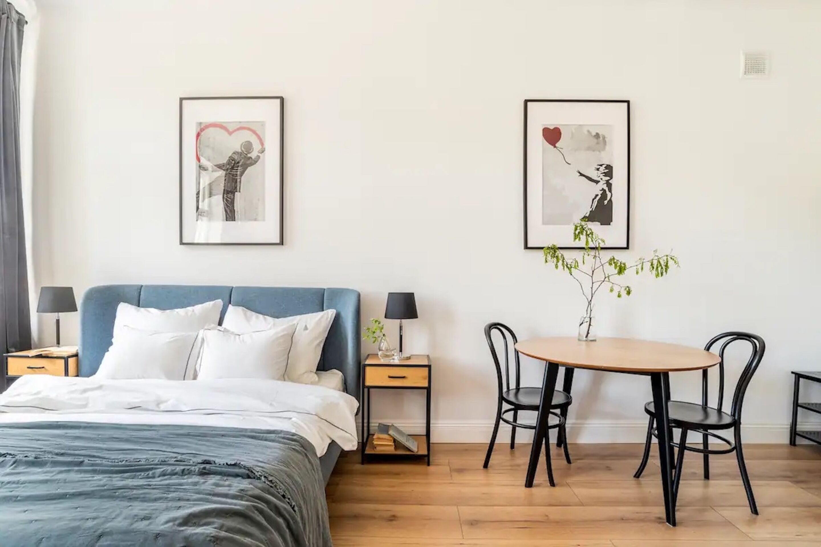 Een slaapkamer met een bed, nachtkastjes, een bureau met stoelen en twee ingelijste kunstwerken aan de muur.