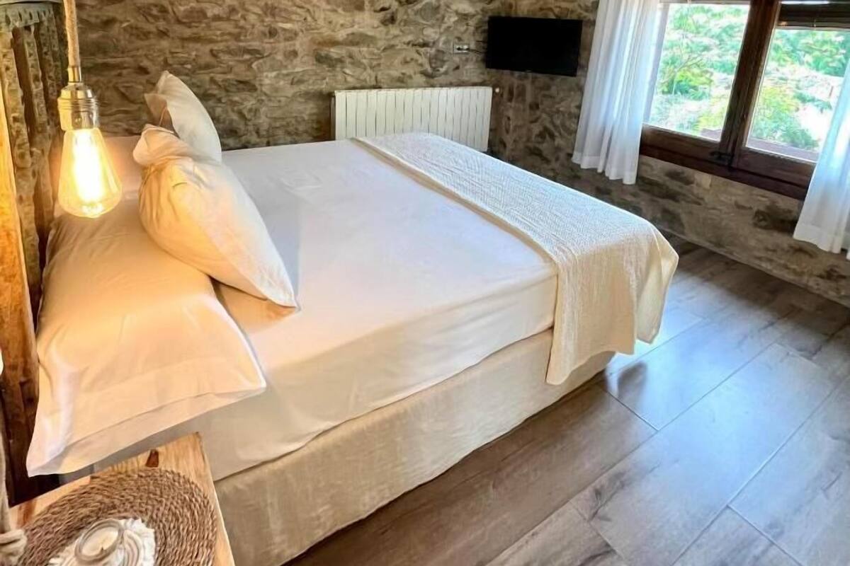 Een slaapkamer met een stenen muur, een bed met witte lakens, een plafondventilator en een raam met gordijnen.