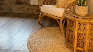 Une chaise en osier avec un coussin et un tapis tissé sur le sol, une petite table en bois avec une plante, et un mur en pierre.