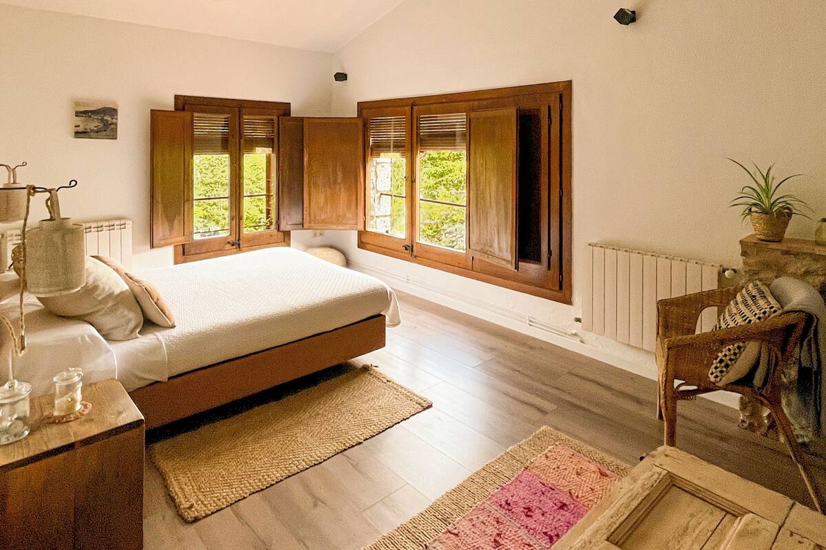 Een slaapkamer met een bed, houten luiken, een radiator, een houten nachtkastje, een stoel en een potplant.