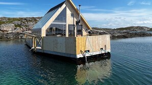 Una casa flotante de madera con techo triangular, ubicada sobre aguas tranquilas con una costa rocosa al fondo.