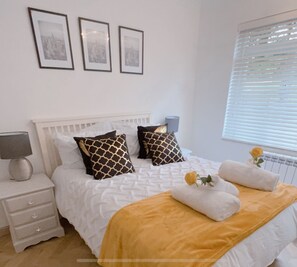 A neatly arranged bedroom with a white bed, patterned pillows, a bedside lamp, and a window with blinds. - Beautiful 3 Bedroom Apartment in Bournemouth (Bournemouth)