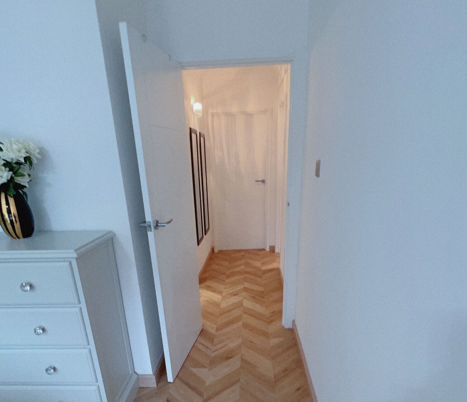 A narrow hallway with a wooden herringbone floor, white walls, and a white door at the end.
